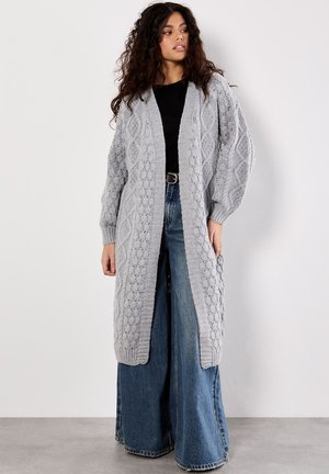 Cardigan lungo aperto in maglia grigia chiara con motivi a rombi e trecce, con polsini a coste, abbinato a jeans in denim blu a gamba larga.