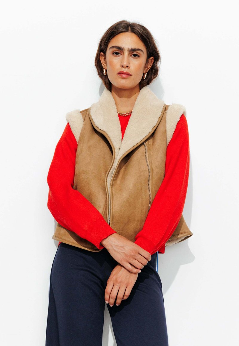Gilet en daim marron avec col en fausse fourrure beige ; fermeture éclair, design sans manches, associé à une chemise à manches longues rouge.