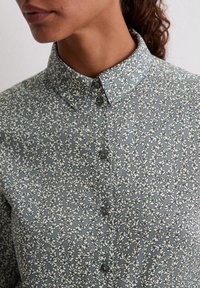 Koszula na guziki w kwiatowy wzór, wykonana z niebiesko-szarej tkaniny z białym kwiatowym motywem, z klasycznym kołnierzem i małymi ciemnymi guzikami.