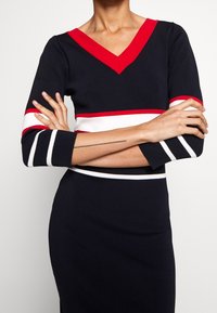 Granatowa sukienka z dekoltem w serek i czerwonym akcentem. Ma białe i czerwone poziome paski przy mankietach i w pasie. Miękki, dopasowany materiał.