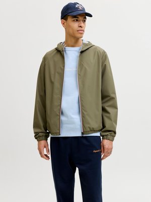 Olíva zöld kapucnis dzsekit viselő férfi világoskék pulóverrel, tengerészkék melegítőnadrággal és tengerészkék sapkával, jobbra néz egy egyszínű háttér előtt.