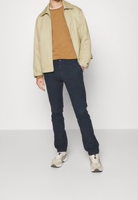 Beige ytterjacka, brun tröja, mörkblå byxor och beige sneakers. Enkel design med texturerad tyg och en avslappnad passform.