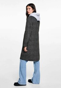 Langer karierter Mantel in Schwarz und Grau, aus einem Wollmix, mit grauem Kapuzenbereich. Kombiniert mit hellblauen weit geschnittenen Jeans und schwarzem Schuhwerk.