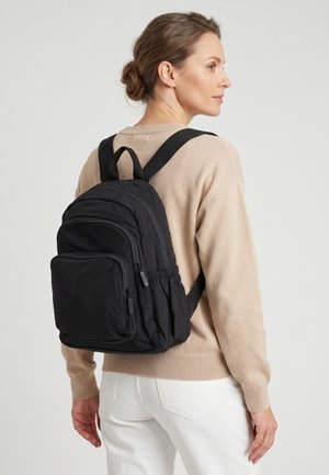 Vrouw met een beige trui en witte broek, die een zwarte rugzak met meerdere ritssluitingvakken draagt, kijkt naar rechts tegen een effen achtergrond.