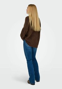 Brun oversized sweater med løs pasform, parret med blå flagrende jeans. Modellen har langt hår og ses fra siden og bagfra.