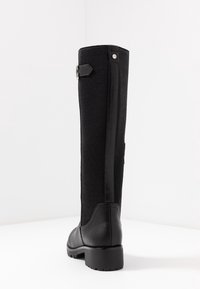 Stivale alto in pelle e tessuto nero con tacco a blocco, cinturino con fibbia vicino alla parte superiore e suola texture, mostrato da dietro su superficie bianca.