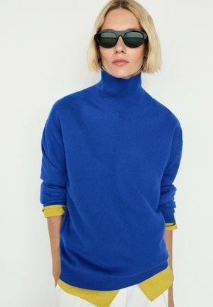 Donna con capelli corti biondi che indossa occhiali da sole neri, un maglione a collo alto blu sopra una camicia gialla e pantaloni bianchi su sfondo uniforme.