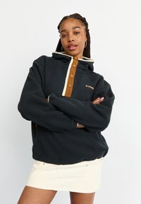 Γυναίκα με πλεξούδες που φοράει μαύρη φλις μπλούζα με καστανά κουμπιά και κρεμ φούστα, στέκεται με τα χέρια σταυρωμένα.