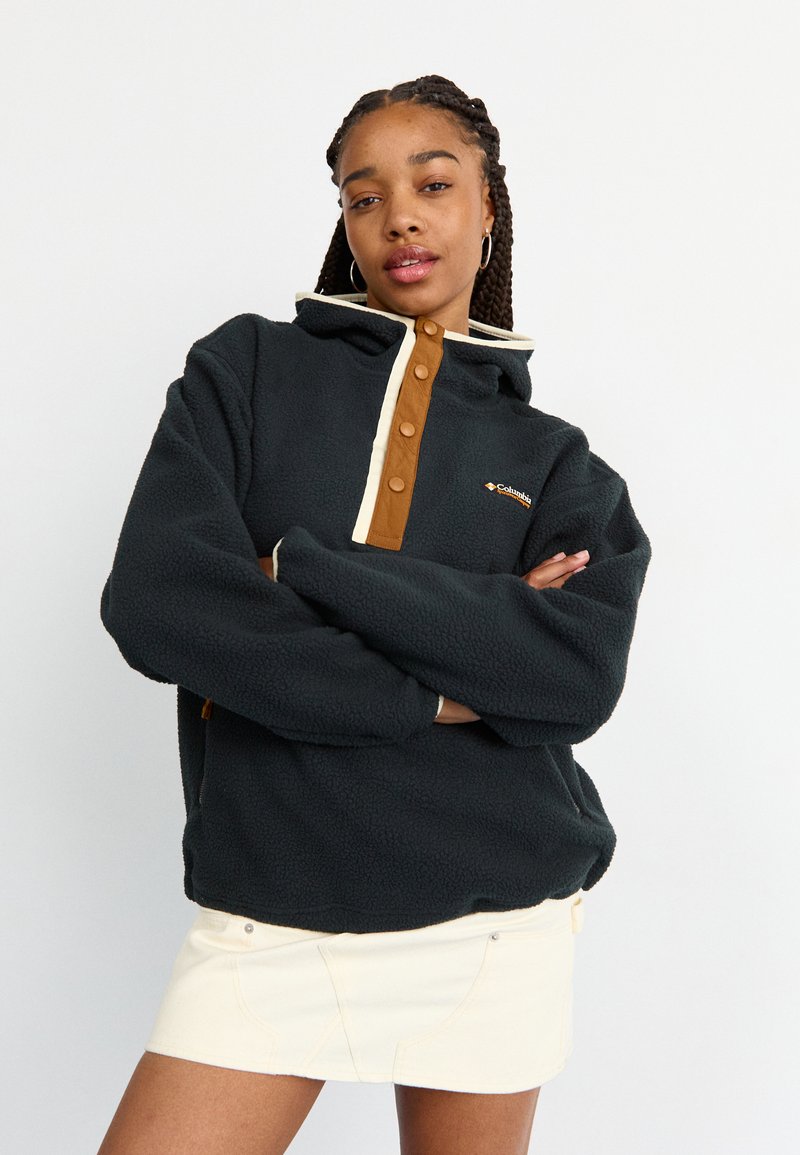 Γυναίκα με πλεξούδες που φοράει μαύρη φλις μπλούζα με καστανά κουμπιά και κρεμ φούστα, στέκεται με τα χέρια σταυρωμένα.