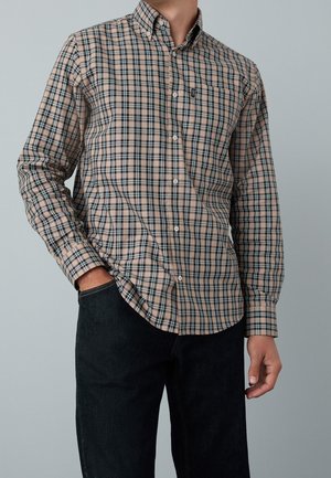 Homme portant une chemise à carreaux beige et noire boutonnée avec une poche avant et un jean foncé, main dans la poche, sur un fond gris clair uni.