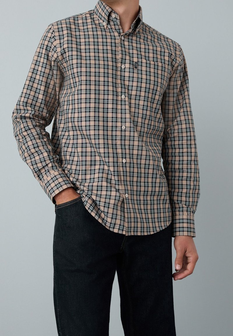 Homme portant une chemise à carreaux beige et noire boutonnée avec une poche avant et un jean foncé, main dans la poche, sur un fond gris clair uni.