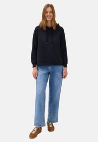 Kvinde står op, iført en sort hættetrøje, lyseblå brede jeans og brune sneakers, smilende mod en ensfarvet hvid baggrund.