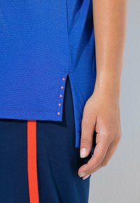 Detail ruky vedle modrého texturovaného lemu košile s malými oranžovými tečkami a tmavě modrých kalhot s vertikálním oranžovým pruhem.