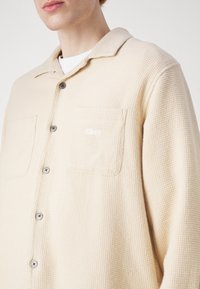 Beige strukturerad skjortjacka med krage, två fickor och bruna knappar; har "Obey" broderat i vitt på vänster ficka.