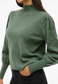 Maglione verde con un colletto alto, maniche a sbuffo texture e polsini a costine. Realizzato in materiale morbido con una finitura liscia.