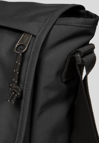 Sac messager noir en tissu texturé, fermeture éclair et bandoulière réglable. Les caractéristiques notables incluent un verrou décoratif et des coutures renforcées.