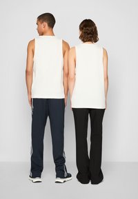 Dos personas están de espaldas, vistiendo camisetas sin mangas de color blanco roto. Una lleva pantalones azul marino con rayas blancas; la otra usa pantalones negros.