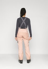 Calças de ski impermeáveis cor-de-rosa com suspensórios cinzentos ajustáveis, bolsos laterais com fecho, e fundos reforçados, usadas sobre uma camisola de manga longa.