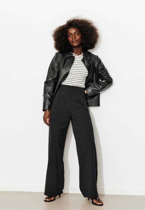 Femme aux cheveux bouclés portant une veste en cuir noire par-dessus un top rayé blanc et noir, un pantalon noir ample et des talons ouverts noirs.