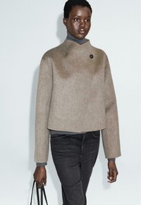 Brun cropped uldjakke med høj krave og knapdetalje, parret med en grå turtle neck og sorte jeans, som holdes med en sort taske.