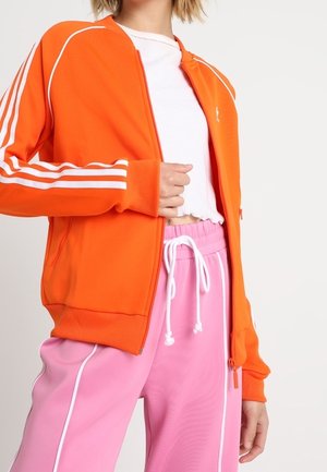 Personne portant une veste zippée orange vif avec des bandes blanches sur les manches, un crop top blanc et un pantalon rose à cordon avec passepoil blanc.