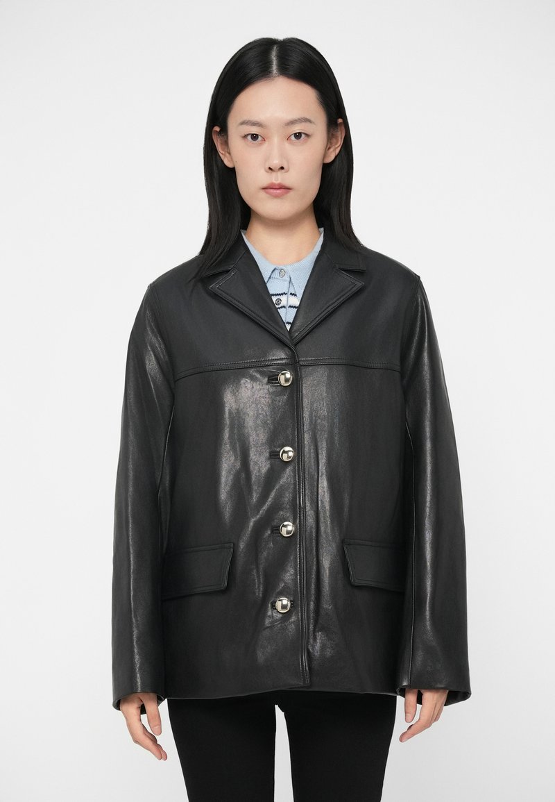 Γυναίκα με ίσια μαύρα μαλλιά φοράει μια μαύρη δερμάτινη �ζακέτα με κουμπιά πάνω από ένα ανοιχτό μπλε πουκάμισο, στέκεται σε λευκό φόντο.