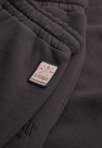 Close-up van donkergrijze stof met elastische manchetten en een rechthoekig Superdry-label met gekruiste golfclubs en de tekst "1954".