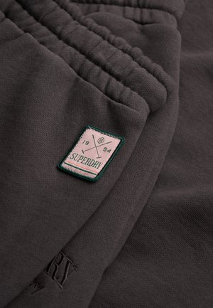 Gros plan sur un tissu gris foncé avec un poignet élastique et un patch rectangulaire Superdry arborant des clubs de golf croisés et le texte "1954".