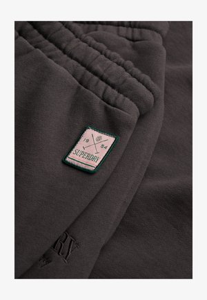 Nærbillede af mørkegrå stof med elastisk manchet og rektangulær Superdry-patch med krydsede golfkøller og teksten "1954".