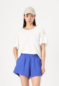 Lys beige caps, løs hvit kortermet skjorte med myk tekstur, og livlige blå shorts med elastisk midje og side lommer.