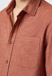 Ruskivärinen nappikauluspaita, jossa on teksturoitu kangas, terävä kaulus ja yksi tasku vasemmassa rinnassa. Napit ovat pyöreät ja ruskeat.