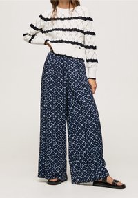Biały sweter dzianinowy w czarne paski, w połączeniu z niebieskimi spodniami typu wide-leg z geometrycznym wzorem. Całość dopełniają czarne płaskie sandały.