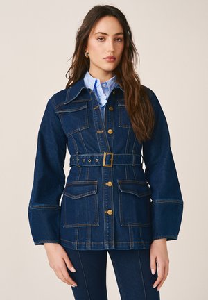 Junge Frau trägt eine dunkelblaue, mit Gürtel versehene Denimjacke mit großen Taschen über einem hellblauen Hemd mit Kragen, steht vor einem schlichten Hintergrund.