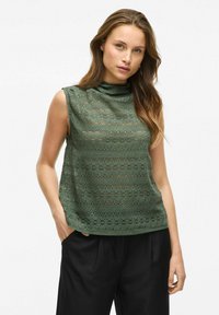 Groen mouwloos topje van kant met een opstaande kraag, voorzien van bloemenpatronen en een losse pasvorm. Gekleed met zwarte wijde broek.