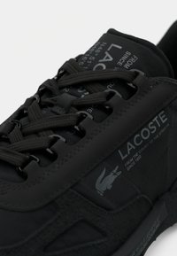Pantof de sport negru din material texturat, cu șireturi, detalii de marcă și un logo distinctiv pe lateral.