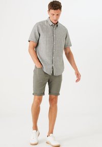 Camisa de manga corta de color gris claro con cierre de botones, combinada con pantalones cortos oliva arremangados. Zapatillas blancas completan el conjunto.