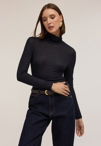 Top negru cu guler înalt, cu un design ajustat, mâneci lungi și o textură ușor transparentă, asortat cu blugi închiși la culoare, cu picioare largi și o curea maro.