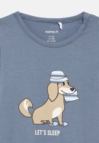 Camisola azul para bebé com um cachorro a sorrir, usando um chapéu de dormir às riscas, chinelos, segurando um travesseiro, e texto "VAMOS DORMIR" abaixo.