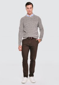 Muž stojí, má na sobě šedý svetr s kulatým výstřihem, světle modrou košili s límečkem, hnědé kalhoty, bílé tenisky a na levém zápěstí náramkové hodinky.