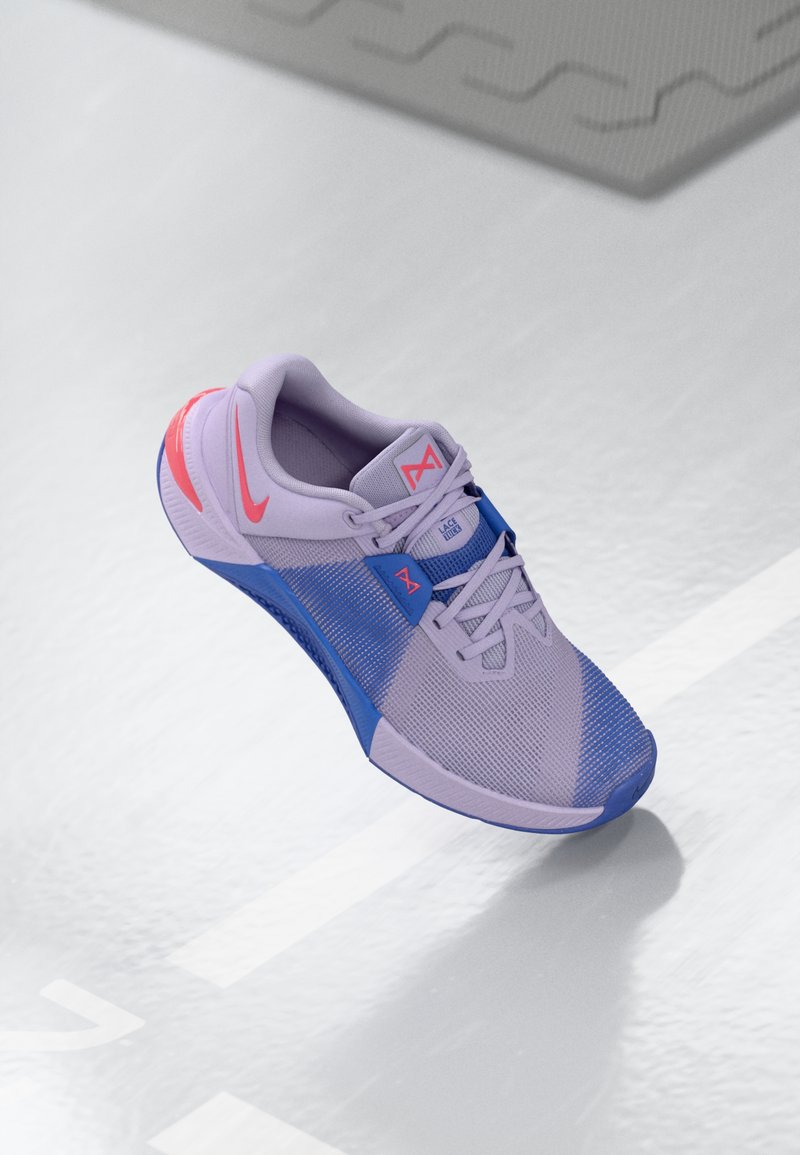 Pantoflă sport violet cu accente albastre, material din plasă și talpă texturată. Include un logo Nike roz și un design cu șireturi pentru o fixare sigură.