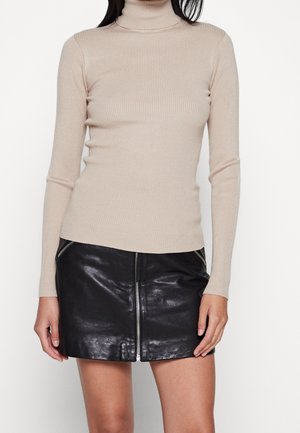 Femme portant un pull à col roulé côtelé beige et une mini-jupe en cuir noire avec fermeture éclair à l'avant et zips aux poches.
