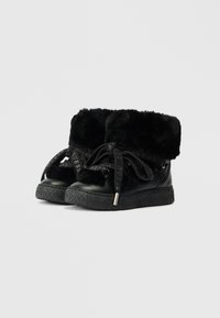 Black ankle boots featuring a faux fur cuff, textured rubber sole, and decorative laces with branded accents. Smooth leather upper.