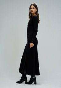 Langes schwarzes Strickkleid mit langen Ärmeln, versehen mit einem seitlichen Schlitz. Kombiniert mit schwarzen, Absatz-Ankle Boots. Glatte Textur und taillierter Schnitt.