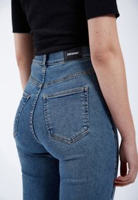Högmidjade blå denimjeans med en åtsittande form, synliga sömmar och två bakfickor; har en svart varumärkesetikett på midjebandet.