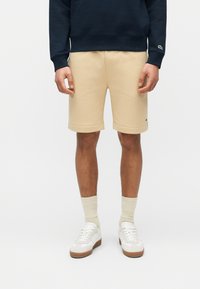 Άνδρας που φοράει μπεζ σορτς μέχρι το γόνατο, κρεμ κάλτσες, λευκά αθλητικά παπούτσια και μπλε σκούρο φούτερ, στέκεται μπροστά σε απλό φόντο.