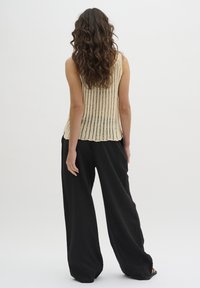 Nainen, jolla on pitkät kiharat hiukset, pukeutuneena olkaimeton beige virkattu toppi ja löysät mustat housut, seisoo selkä kameraan yksinkertaisella taustalla.