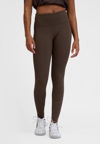 Bruna leggings gjorda av stretchmaterial, med ett högt midjeband och slät yta. Bärs med vita träningsskor.