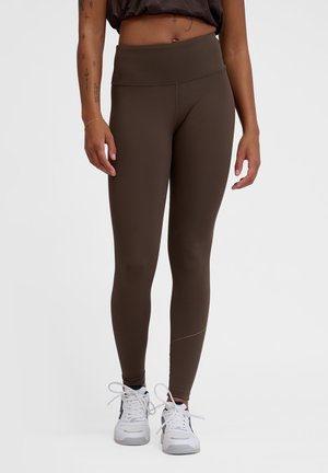 Bruine leggings van rekbaar materiaal, met een hoge tailleband en een gladde textuur. Gemaakt in combinatie met witte sportschoenen.