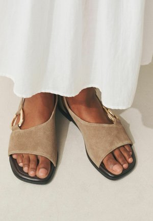 Dunkelhäutige Füße, die beige Wildleder-Sandalen mit offenen Zehen und goldenen Schnallenakzenten tragen, stehen auf einem hellen Boden unter eines weißen Kleidungsstücks.