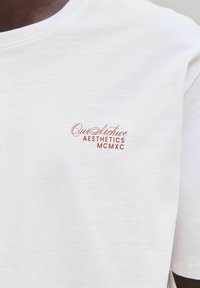 Hvid t-shirt i bomuld med en lille, broderet logo i rødbrun farve, der siger "Our Archive AESTHETICS MCMXC." Teksten er placeret på brystet.